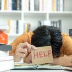 woman with her head on her work desk holding a sign that says help because she doesnt know how to cope with high functioning anxiety