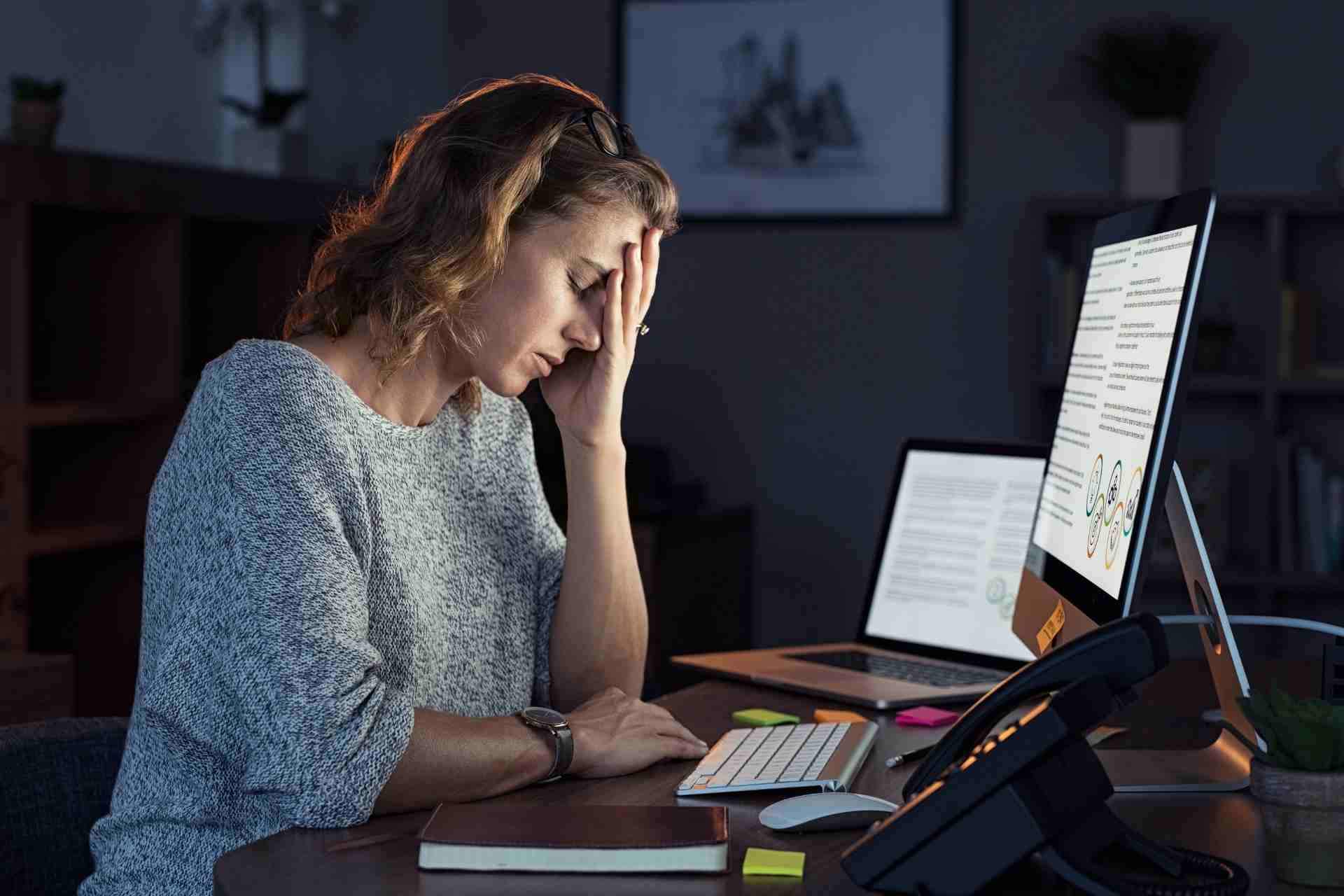 Woman experiencing mental health burnout at work while sitting at her desk with her hand on her forehead during late-night office hours.