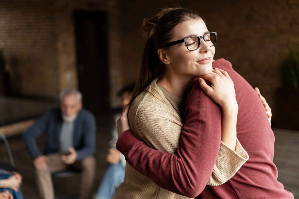 Woman with glasses embracing another person in a comforting hug during a supportive group gathering.